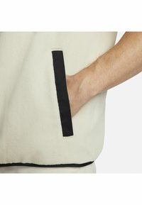 Chaleco de forro polar de color crema claro con un ribete negro en contraste y un bolsillo. La textura parece suave, con detalles de diseño mínimos.