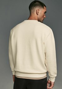 Cremefarvet sweatshirt med tekstureret stof, ribbede mansjetter og kant samt en afslappet pasform. Bagsiden viser et enkelt, rent design.