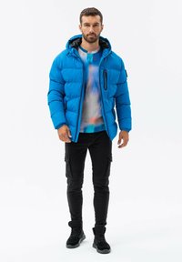 Modrá zateplená bunda s kapucí, s předním zipem a boční kapsou. Nositá přes tričko s mnohobarevným tie-dye vzorem, v kombinaci s černými cargo kalhotami.