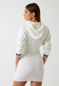 Vestido de punto blanco con capucha, que presenta detalles a rayas en verde oscuro, rojo y blanco en las mangas. Textura suave y diseño ajustado.