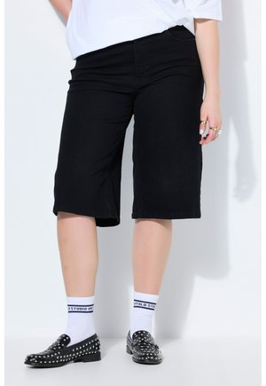 Osoba nosząca czarne krótkie spodenki do kolan, białe skarpetki do kostek z czarnym napisem, czarne mokasyny z ćwiekami oraz białą, włożoną do środka koszulę.