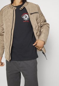 Béžová prošívaná bunda s klopou na patentky a zipovou kapsou, nošená přes černé tričko s červeným a bílým grafickým designem, doplněná o šedé kalhoty.