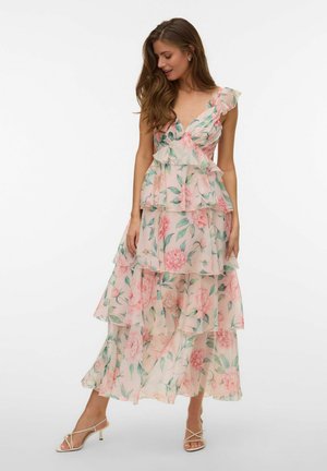 Femme portant une robe maxi sans manches à volants avec des fleurs roses et des feuilles vertes, associée à des talons beiges à brides.