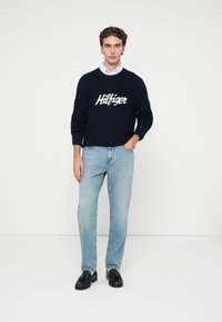 Granatowy sweter dzianinowy z logo "Hilfiger" na przodzie, w zestawie z jasnoniebieskimi dżinsami prostymi i czarnymi mokasynami. Gładkie tło.
