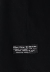 Etiqueta de tela negra con texto blanco que dice "ESCAPAR DE / LO DESCONOCIDO" y detalles adicionales de la marca. Textura suave, forma rectangular.