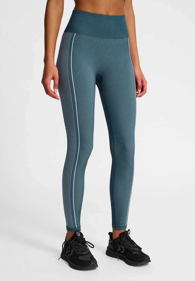 Główne legginsy sportowe w kolorze turkusowym z wysokim stanem, o fakturze ryflowanej i białych pionowych paskach po bokach. W połączeniu z czarnymi butami sportowymi.