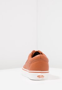 Sneaker en cuir marron avec bout rond, surface texturée, semelle intermédiaire blanche et accent de logo sur le talon. Design minimaliste, silhouette basse.