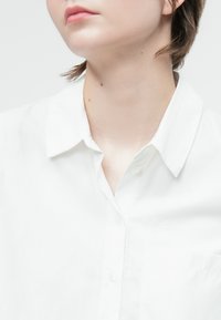 Camicia bianca a collare realizzata in tessuto leggero, con chiusura a bottoni sul davanti e una piccola tasca sul lato sinistro.