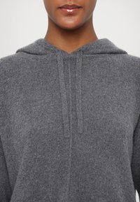 Gros plan d’une personne portant un sweat à capuche gris tricoté doux avec des cordons, montrant les épaules et les lèvres sur un fond uni.