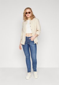 Beige linnen knappskjorte over en hvit cropped topp, parret med blå skinny jeans og hvite sneakers. Solbriller fullfører stilen.