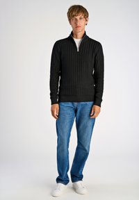 Schwarzer Reißverschluss-Pullover mit strukturiertem Rippstrickdesign, kombiniert mit hellblauen Jeans und weißen Sneakern. Einfaches und lässiges Outfit.