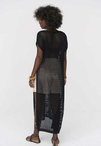Zwarte gehaakte maxi cover-up met open gebreid ontwerp, korte mouwen en zijsplitten, gedragen over een aansluitende outfit. Textuur en lichtgewicht.