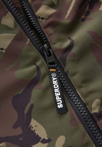 Chaqueta con cremallera y diseño de camuflaje, con una cremallera negra, tela texturizada y una etiqueta de Superdry. Predominantemente verde con patrones marrones y beige.