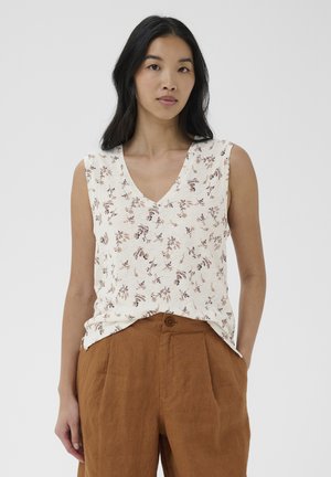 Femme portant un haut blanc sans manches à col en V avec un motif floral et un pantalon marron taille haute, sur un fond uni.