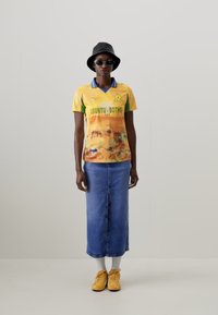 T-shirt grafico giallo e verde con "UBUNTU BOTHO", abbinato a una gonna di denim blu. Gli accessori includono un cappello a secchiello nero e occhiali da sole. Sneakers gialle.