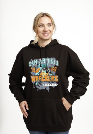 Femme souriante portant un sweat à capuche noir avec le texte « San Antonio Wreckers Baseball 1997 » et un graphisme futuriste d'un joueur de baseball à l'avant.