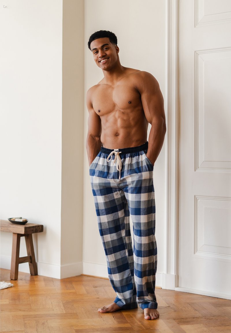 Een shirtloze man die glimlacht, staande op blote voeten binnen, gekleed in blauwe en witte geruite pyjamabroek met zijn handen in de zakken.