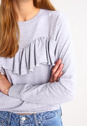 Frau trägt ein hellgraues Langarmshirt mit Rüschendetail über der Brust und blaue Jeans, die Arme verschränkt.
