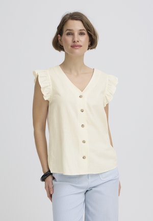 Žena s krátkymi hnedými vlasmi, oblečená v krémovej blúzke bez rukávov s volánikmi a gombíkmi a svetlomodrých nohaviciach s prúžkami na bielom pozadí.