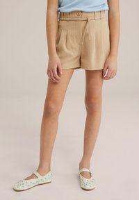 Beige skräddarsydda shorts med veck och bälte, i kombination med vita Mary Jane-skor dekorerade med metalliska nitar. Blå ribbad topp synlig.