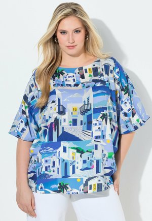 Donna bionda che indossa una maglia blu morbida con stampe bianche di edifici in stile mediterraneo, abbinata a pantaloni bianchi, su sfondo uniforme.