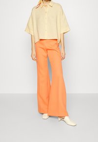 Kvinde iført en løs, cropped beige knap-ned skjorte, lyse orange bredbenede bukser, cremefarvede sko med firkantet tå og et gult tørklæde i håret.