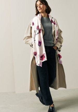 Femme portant un trench-coat beige, un pull gris, un pantalon noir, des chaussures noires et une écharpe blanche à imprimé floral rose, debout les mains dans les poches.