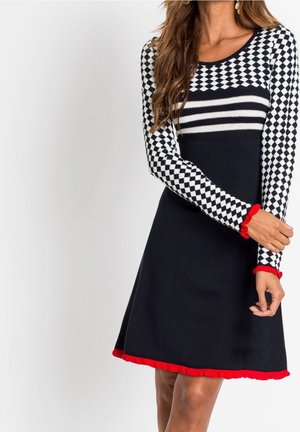Donna che indossa un vestito nero con maniche e parte superiore a quadretti bianco e nero, righe orizzontali e bordi rossi su polsini e orlo.