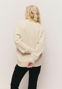 Beige, strukturovaný oversize svetr s vroubkovanými manžetami a lemem, spárovaný s černými kalhotami. Pohled ze zad, modelka si drží ruce za zády.