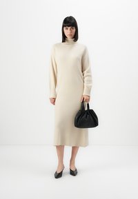 Vestito lungo in maglia beige con collo alto e maniche larghe, abbinato a una borsa nera arricciata e scarpe con tacco nere. Tessuto liscio.