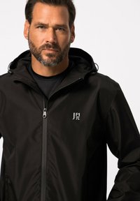 Zwarte waterdichte jas met ritssluiting en capuchon. Heeft een gestructureerd oppervlak en een klein wit logo op de linkerkant van de borst.