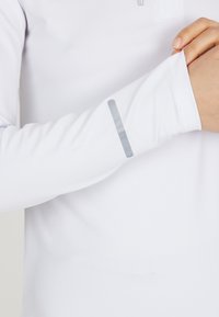 Weißes Langarm-Performance-Shirt mit glatter Textur, ausgestattet mit einem reflektierenden grauen Akzent am Bündchen und flachen Nähten für zusätzlichen Komfort.