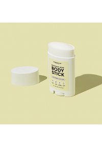 Boîtier stick blanc ouvert étiqueté « Glycolic Acid Body Stick » avec parfum verveine citronnée et menthe sur fond jaune pâle.