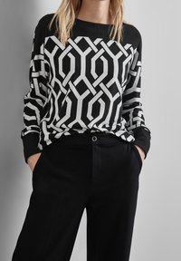 Svart och vit mönstrad tröja med geometrisk design; långa ärmar och ribbade muddar; ihop med svarta byxor med en framficka.