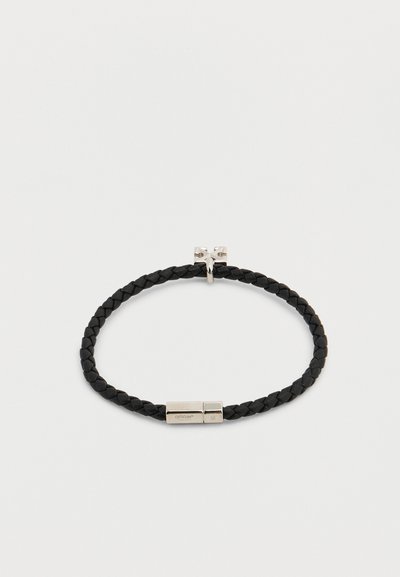 Bracelet en cuir tressé noir avec fermoir magnétique en argent et petit pendentif croix en argent sur fond clair.