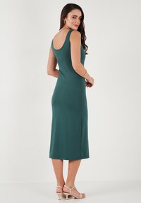 Varren, polvenpituinen vihreä jersey-mekko, jossa on tiukka siluetti. Mekossa on avara selkä ja sivuaukko, ja se on yhdistetty nude-henkisiin remmisandaaleihin.