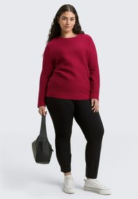 Maronenfarbener strukturierter Pullover mit langen Ärmeln, kombiniert mit schwarzen Slim-Fit-Hosen und weißen Sneakers, während eine schwarze Tragetasche gehalten wird.