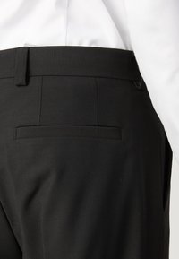 Schwarze Hosen mit glatter Textur, die über eine einzelne Gesäßtasche und ein sauberes Design des Taillenbundes verfügen, teilweise sichtbar über einem weißen Hemd.