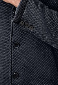 Sort tekstureret jakke med en tætsiddende pasform, detaljeret stikning og fire runde knapper på ærmet. Stoffet har et vævet mønster.
