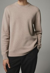 Beżowy sweter dzianinowy z okrągłym dekoltem i ściągaczami, w połączeniu z czarnymi spodniami w paski. Miękka tekstura i luźny krój.