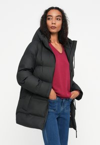 Crni puffer kaput s kapuljačom, s prednjim zatvaračem i elastičnim manžetama. Nosi se preko bordo V-izreza majice i plavih traperica.