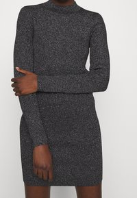 Robe en tissu tricoté noir avec des paillettes métalliques, dotée d'un col haut et de manches longues, ajustée à la silhouette et d'une longueur mi-cuisse.