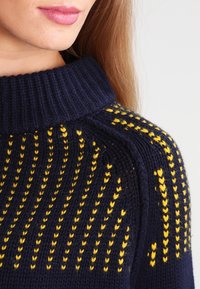 Il maglione a maglia blu navy presenta un colletto a ribalta con un motivo a zigzag giallo, evidenziando il materiale in lana texturizzata e un design aderente.