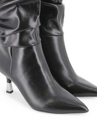 Botas de tobillo de cuero negro con puntera afilada, diseño plegado en la parte superior y tacón cuadrado de metal plateado. Textura suave con pliegues sutiles.