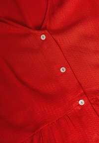 Röd klänning med ett texturerat mönster, med V-ringning, små vita knappar och rynkad tyg vid midjan. Slätt och lätt material.