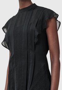 Robe noire avec des panneaux verticaux texturés et des manches volantées portée par une personne à la peau foncée, montrée du cou à la taille sur un fond uni.
