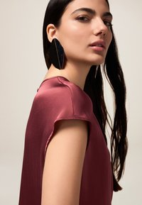Top in satin bordeaux con maniche corte, caratterizzato da un colletto rotondo. Un grande orecchino geometrico nero accentua il look. Texture liscia, design minimale.