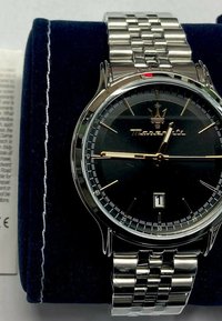 Orologio in acciaio inossidabile argento con quadrante nero rotondo, dettagli dorati e finestra per la data. Attaccato a un bracciale a maglie metalliche. Presentato su un cuscino blu.