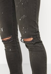 Jeans ajustados negros desgastados con aberturas deshilachadas en las rodillas y detalles de salpicaduras de pintura blanca. Hechos de mezclilla para un ajuste ceñido.