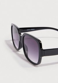 Μαύρα oversized γυαλιά ηλίου με βαθμιαία μοβ φακούς. Διαθέτουν γυαλιστερό πλαστικό σκελετό και παχιά τετράγωνα μπράτσα, με το λογότυπο "ANNA FIELD."